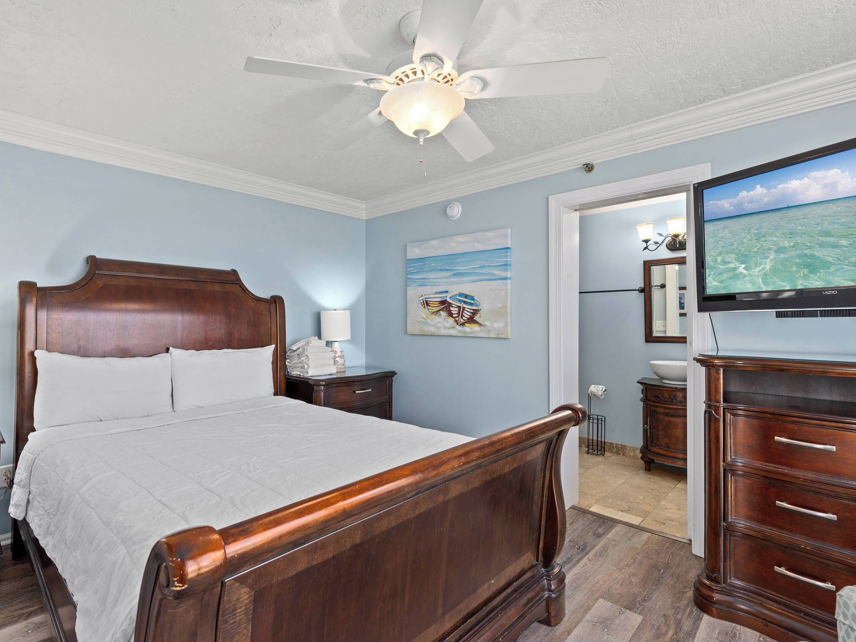 Une chambre à coucher avec un lit en bois, un ventilateur de plafond, un tableau représentant des bateaux sur une plage et un téléviseur posé sur une commode.