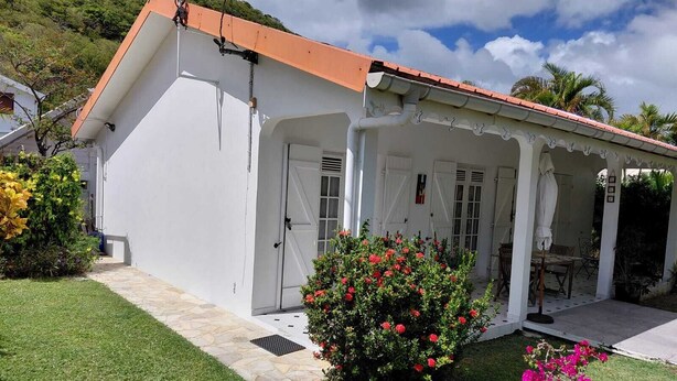A white building with a red roof, a covered porch, and a small garden with red flowers. - Maine-Charming Creole villa at the gateway to the south of Martinique. (sainte Anne)