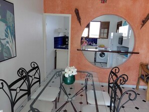 A dining area with a glass table, metal chairs, a vase with flowers, and a kitchen visible through a round mirror. - Maine-Charming Creole villa at the gateway to the south of Martinique. (sainte Anne)
