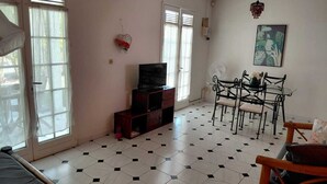 A room with a bed, a TV, a dining table with chairs, a fan, and a window with blinds. - Maine-Charming Creole villa at the gateway to the south of Martinique. (sainte Anne)
