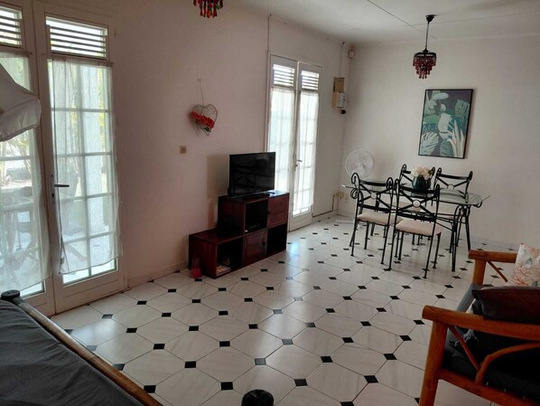 A room with a bed, a TV, a dining table with chairs, a fan, and a window with blinds. - Maine-Charming Creole villa at the gateway to the south of Martinique. (sainte Anne)