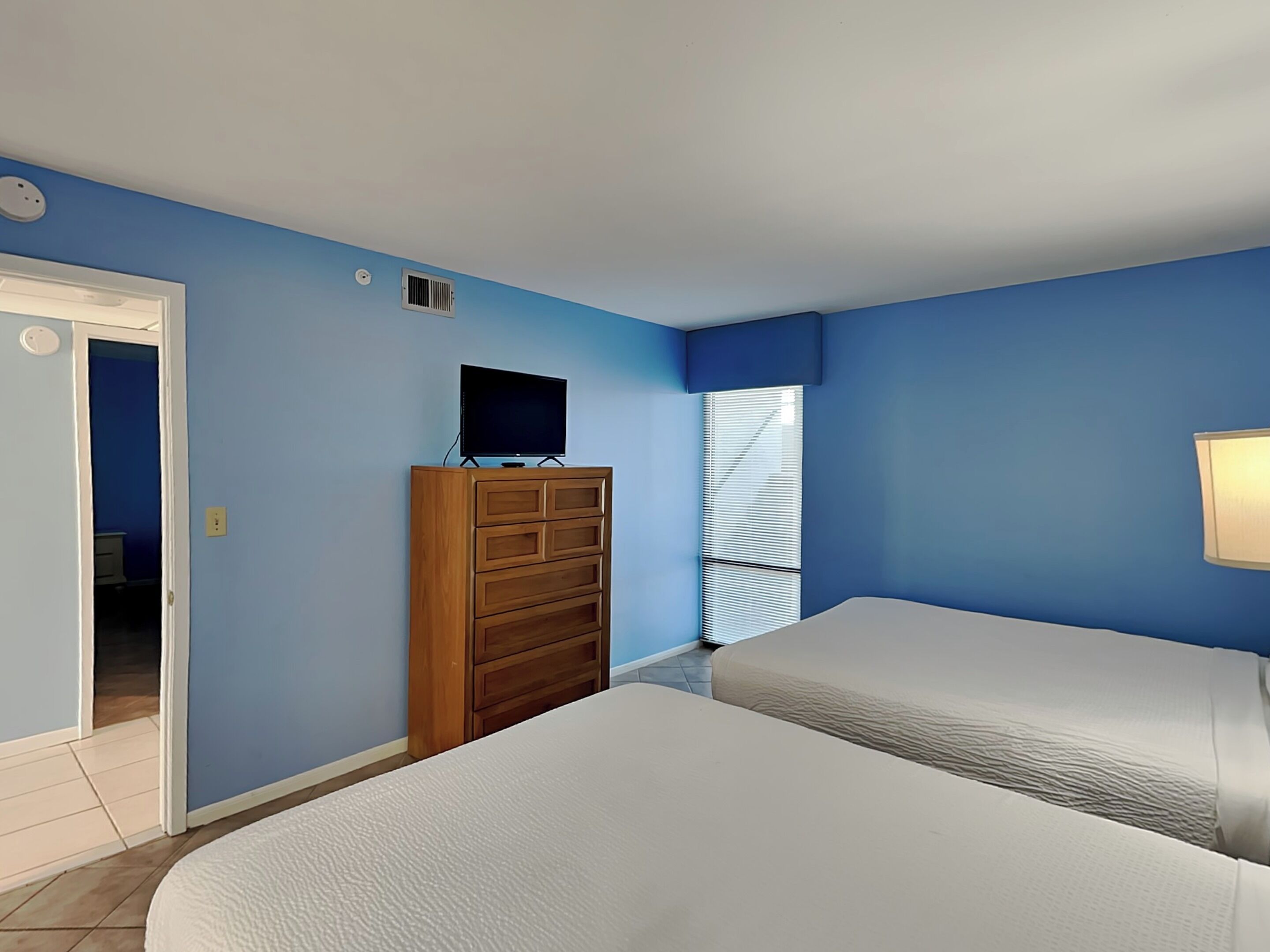 Un dormitorio con dos camas, un aparador de madera, un televisor encima y una ventana con persianas.