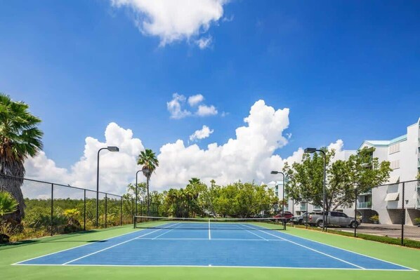 Un court de tennis extérieur doté d’une surface bleue, entouré d’une clôture, de palmiers et de bâtiments en arrière-plan.