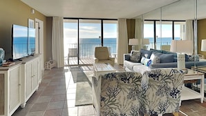 A spacious living room with a large flat-screen TV, a sofa, a chair, and a dining table. The room has a balcony with a view of the ocean.