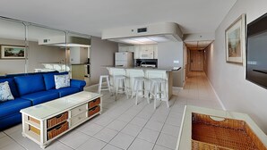 Un salon moderne avec un canapé bleu, une table basse blanche et un coin cuisine doté de tabourets blancs.