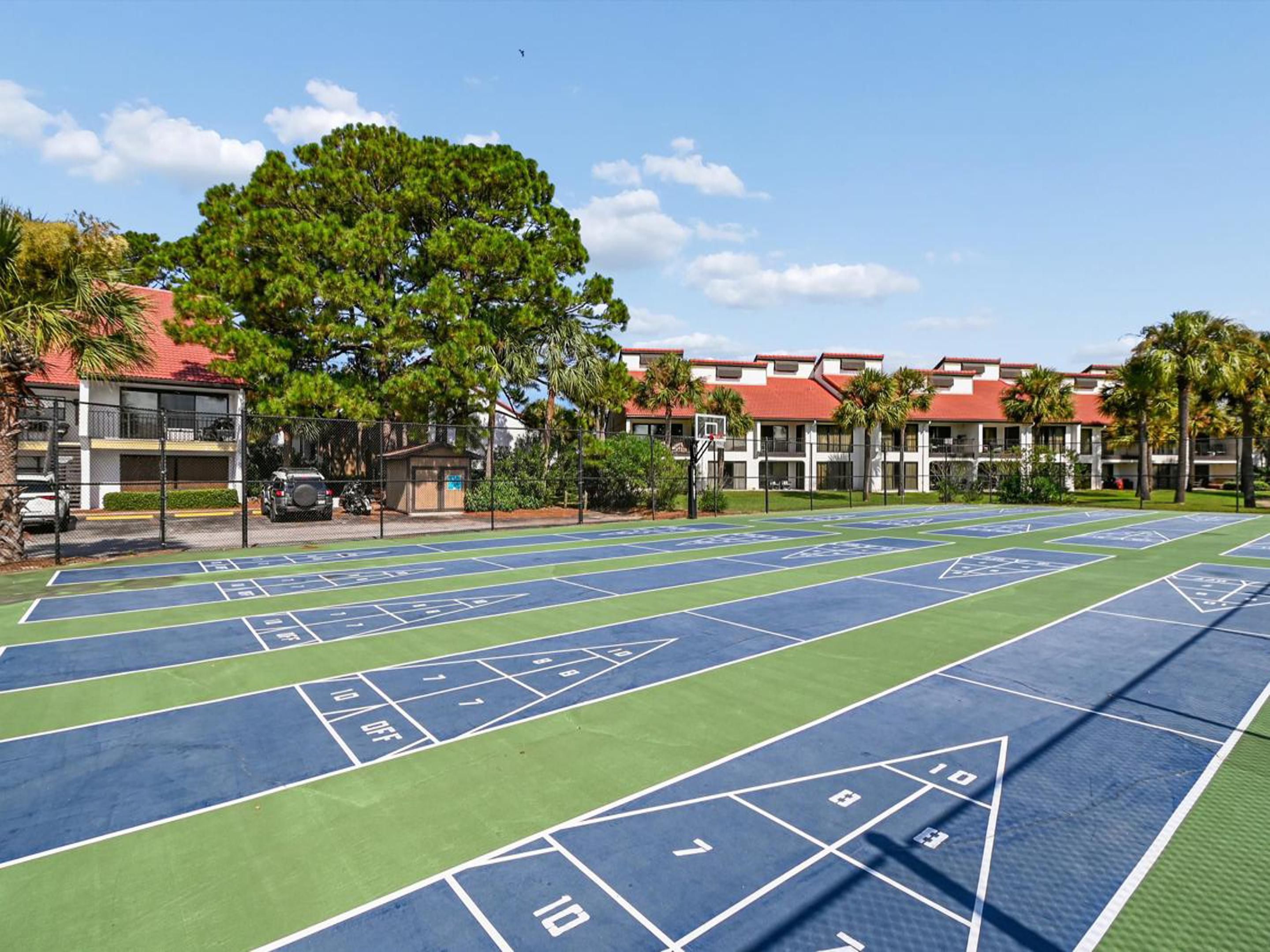 Una cancha de shuffleboard con cielo despejado, rodeada de edificios con techos rojos y palmeras.