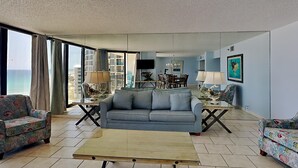 Ein Wohnzimmer mit einer Couch, zwei Sesseln, einem Couchtisch und einem Essbereich mit Fernseher und Tischleuchten.