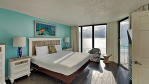 A large bed with a wooden headboard, two bedside tables with lamps, a chair, and a large window with a view of the ocean. - Edgewater Tower III #804 | Beachside Pool + Views! (Panama City Beach)