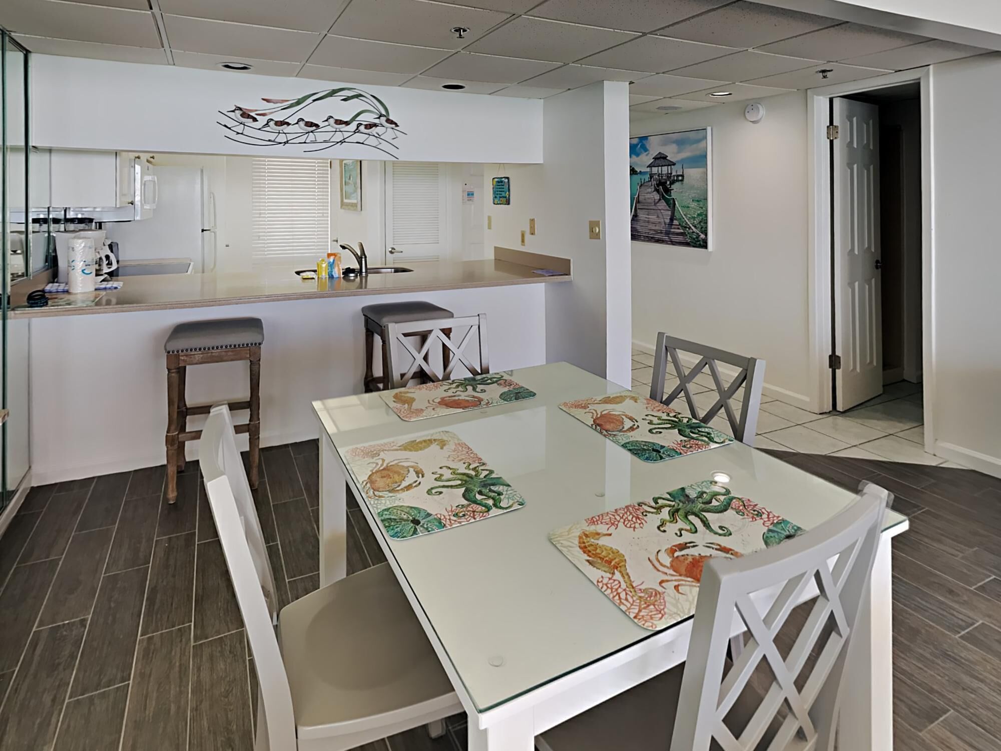 A dining area with a glass table, white chairs, and a kitchen in the background.
