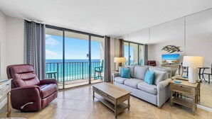 Un salón amplio con balcón y vistas al océano, un sillón de cuero, un sofá con cojines, una mesa de centro de madera y un televisor.