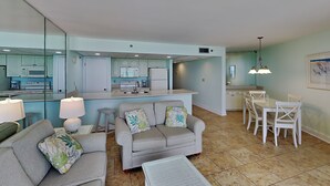 A modern living room with a grey sofa, a coffee table, and a dining area with white chairs and a table. - Edgewater Tower III #910 | Gulf Front + Amenities (Panama City Beach)