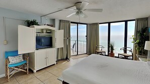 A hotel room with a bed, a TV, a ceiling fan, a balcony with a view of the ocean, and a chair with a cushion. - Edgewater Tower III #911 | Beachfront + Amenities! (Panama City Beach)