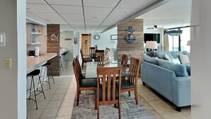A modern living room with a sofa, a dining area with a table and chairs, and a kitchen area with a counter and stools. - Edgewater Tower III #911 | Beachfront + Amenities! (Panama City Beach)