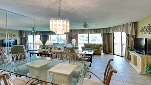 Un salon spacieux avec une table à manger au plateau en verre, des sièges confortables, un lustre et un téléviseur.