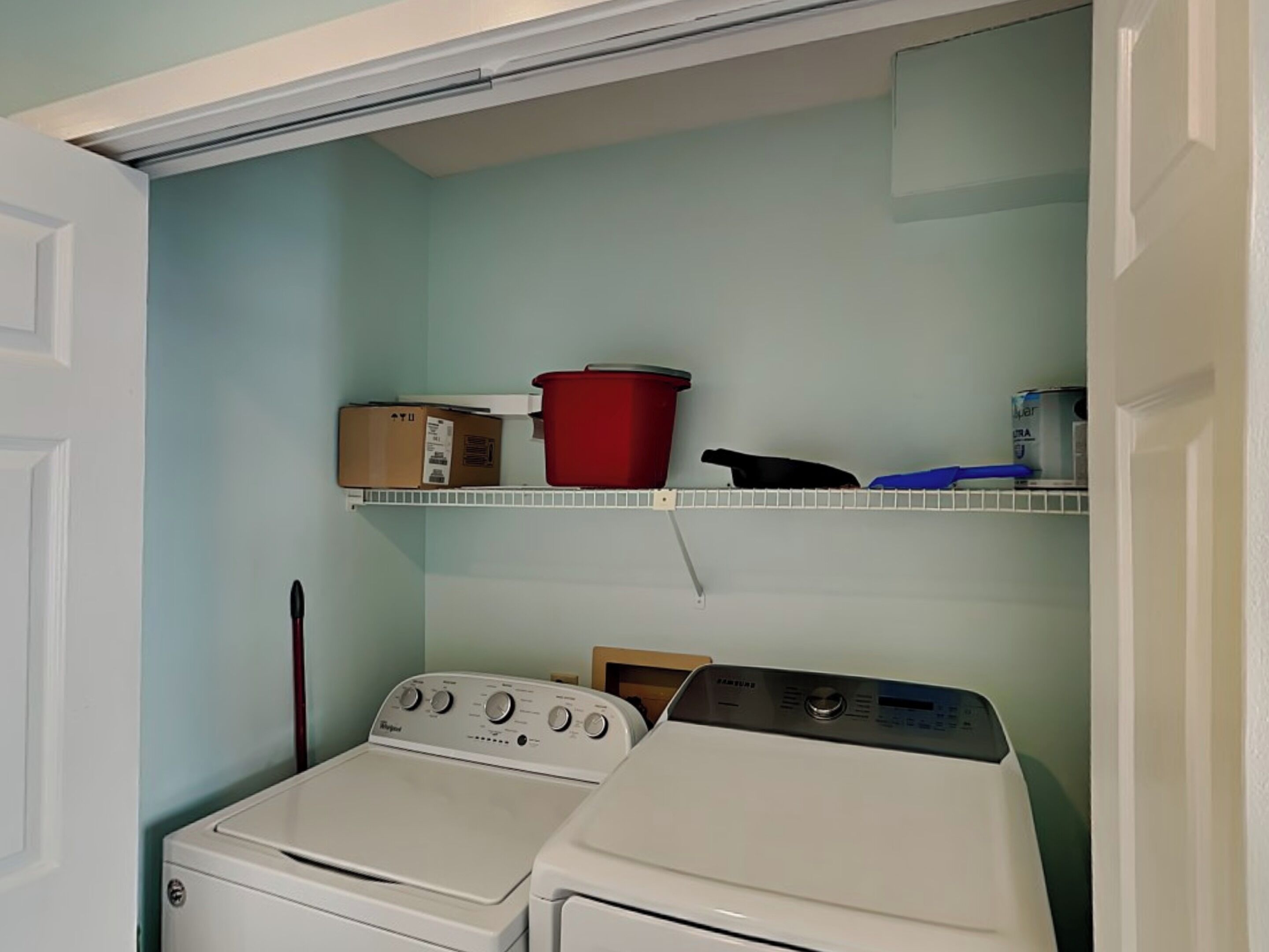 A laundry room with two stacked washers and dryers, a red pot, a black pan, and a blue cleaning brush on a shelf.