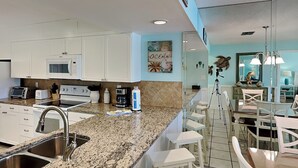 Cocina moderna con armarios blancos, encimeras de granito y pileta de acero inoxidable. Cuenta con una zona de comedor con taburetes blancos y un espejo grande sobre la encimera.