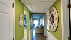 A hallway with green walls, white trim, and a large wall clock. There is a pineapple decoration on the left wall and a window with curtains in the background. - Edgewater Villa #1702 | Golf View and Resort Amenities (Panama City Beach)