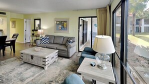 Un salon comprenant un canapé, une table basse et des fauteuils, un coin repas avec une table et des chaises, et une grande fenêtre donnant sur l’extérieur.