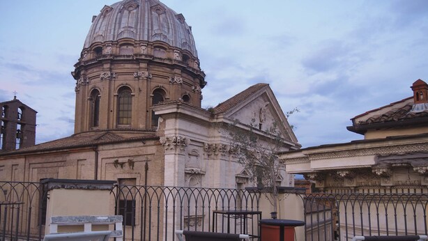 Une terrasse avec deux chaises pliantes blanches et une table, d’où l’on peut admirer un bâtiment historique doté d’un dôme et d’une architecture complexe.