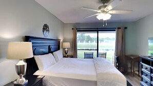 A bedroom with a large bed, two bedside tables with lamps, a ceiling fan, and a view of a golf course through the window. - Edgewater Golf Villa #1908 | The Best Amenities! (Panama City Beach)