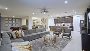 Un salón amplio con un sofá seccional gris, una mesa de centro y un comedor con sillas y una mesa. Se ve una cocina con armarios y electrodomésticos al fondo.