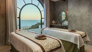 Une chambre avec deux lits, une grande fenêtre donnant sur la mer, un miroir rond et une petite table avec un plateau.