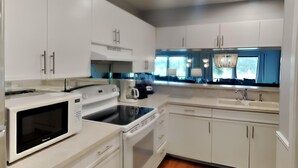 A modern kitchen with white cabinets, a microwave, an oven, a coffee maker, and a window with blue curtains. - Edgewater Villa #1913 | Beach Access, Pools, & Golf! (Panama City Beach)