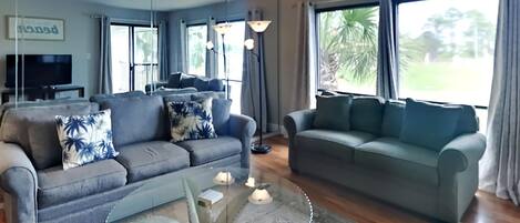 Ein Wohnzimmer mit zwei Sofas, einem Glastisch und einem Fernseher im Hintergrund.