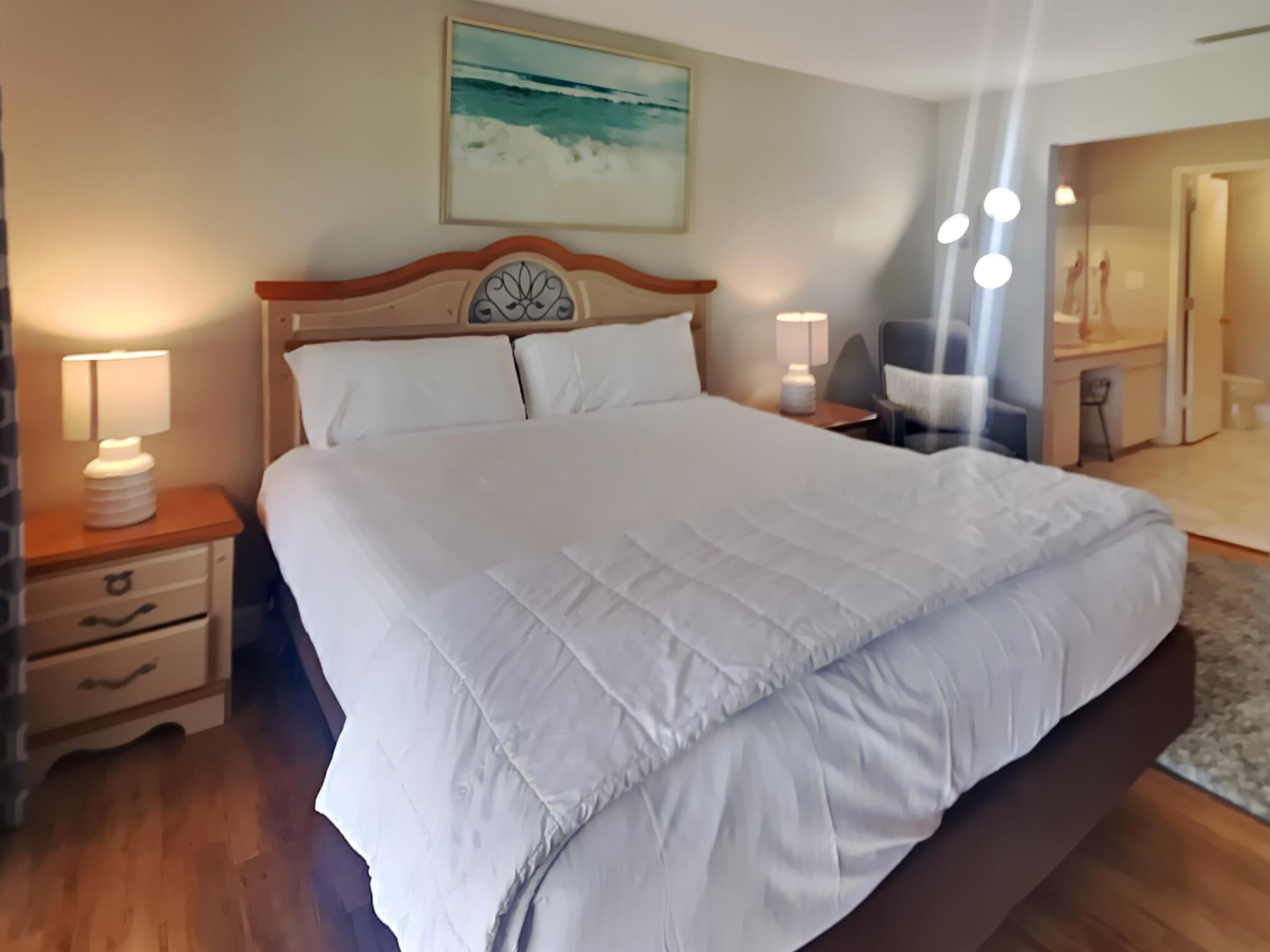 Una cama bien hecha con sábanas blancas, cabecera de madera, mesitas de noche con lámparas y un cuadro con una escena de playa en la pared.