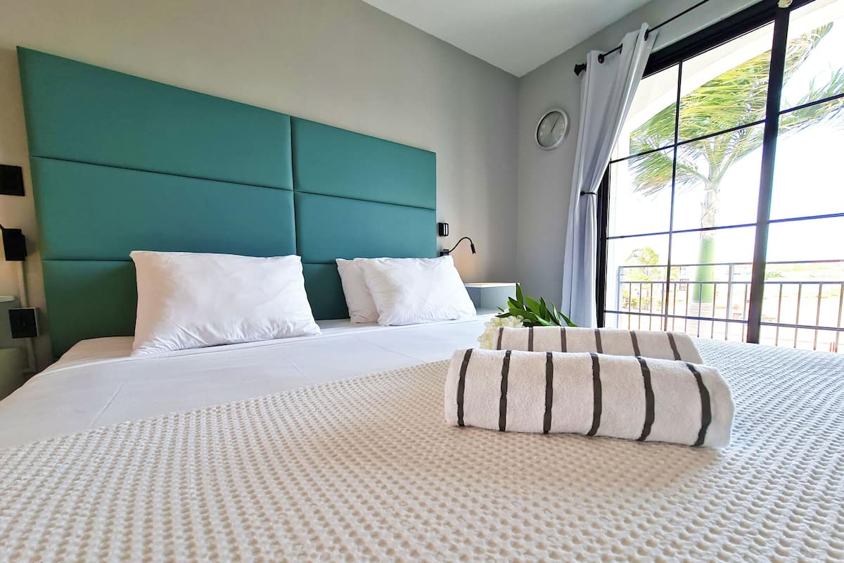 Habitación de hotel con una cama grande, cabecera color turquesa, ropa de cama blanca y vista a palmeras a través de la ventana.