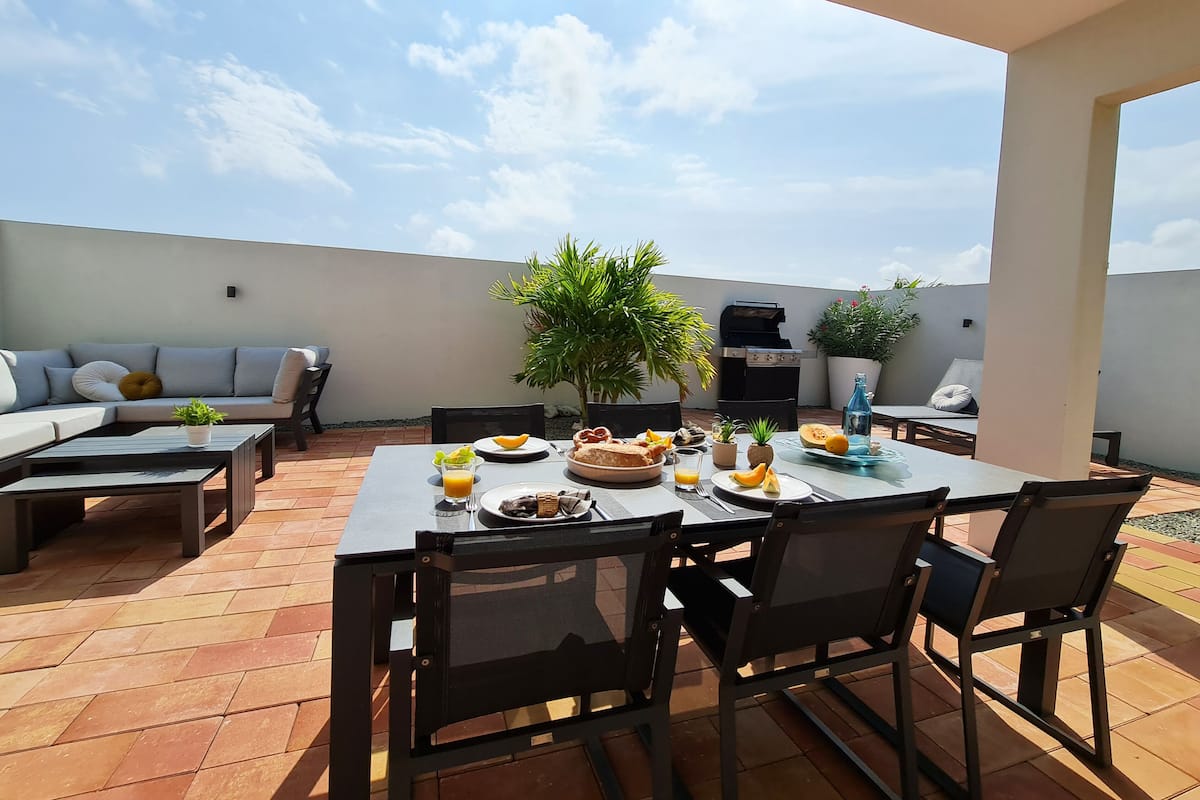 Una terraza en la azotea con una mesa de comedor lista para dos personas, rodeada de sillas, un sofá y una parrilla.