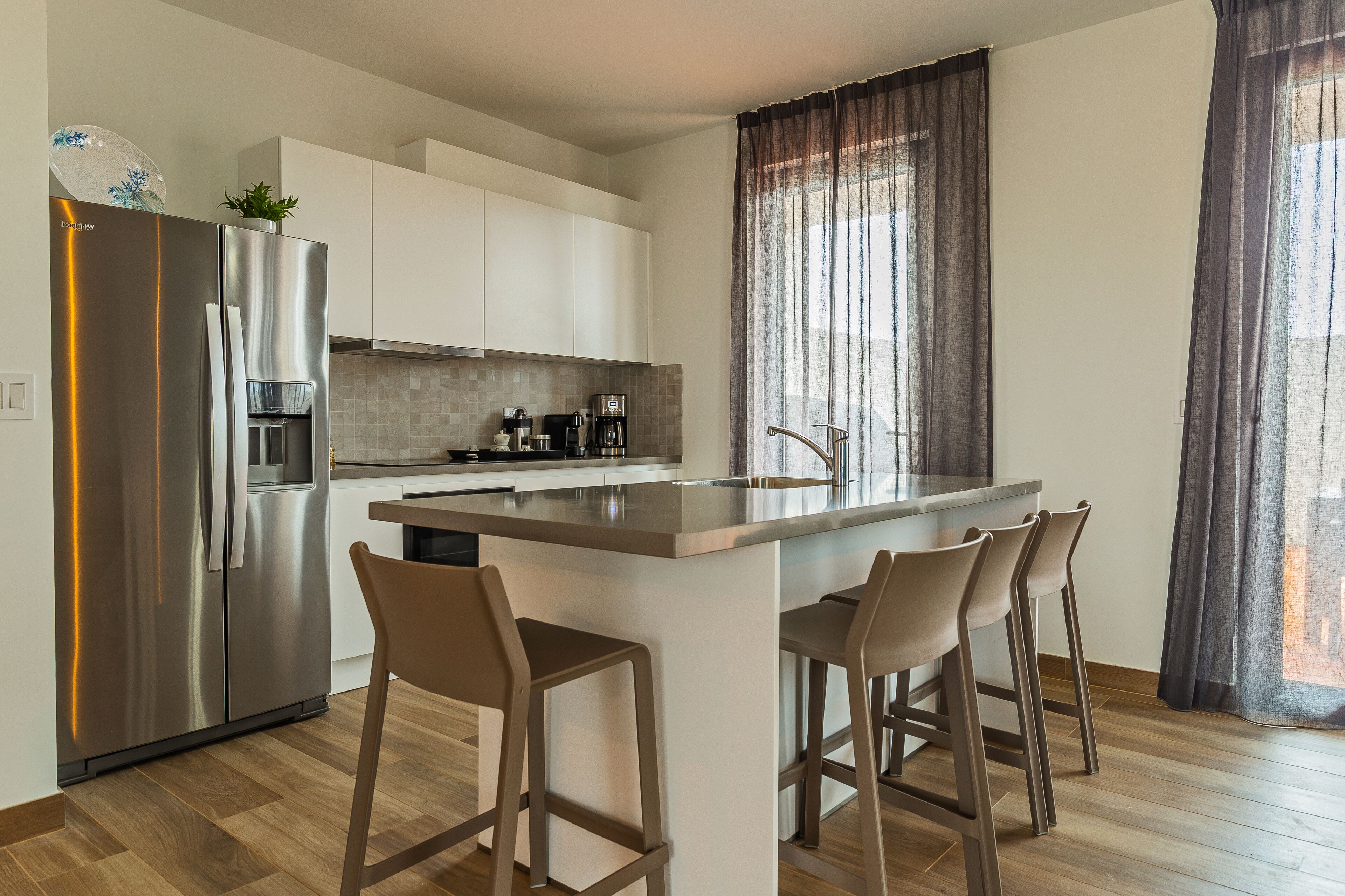 A modern kitchen with a stainless steel refrigerator, a built-in oven, and a coffee maker. There are bar stools at the island and a window with curtains.