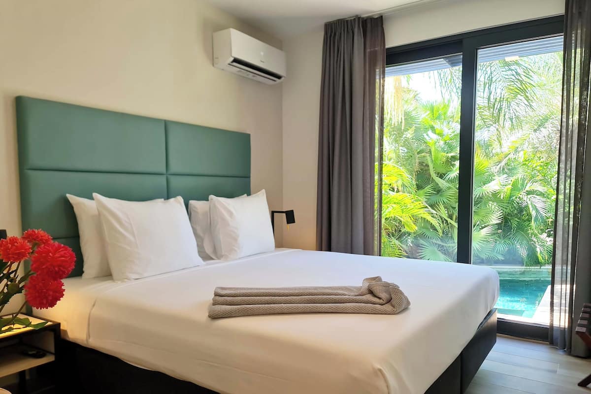 Habitación de hotel con una cama grande, cabecera verde, ropa de cama blanca, una toalla y vista a una piscina a través de una puerta de cristal.