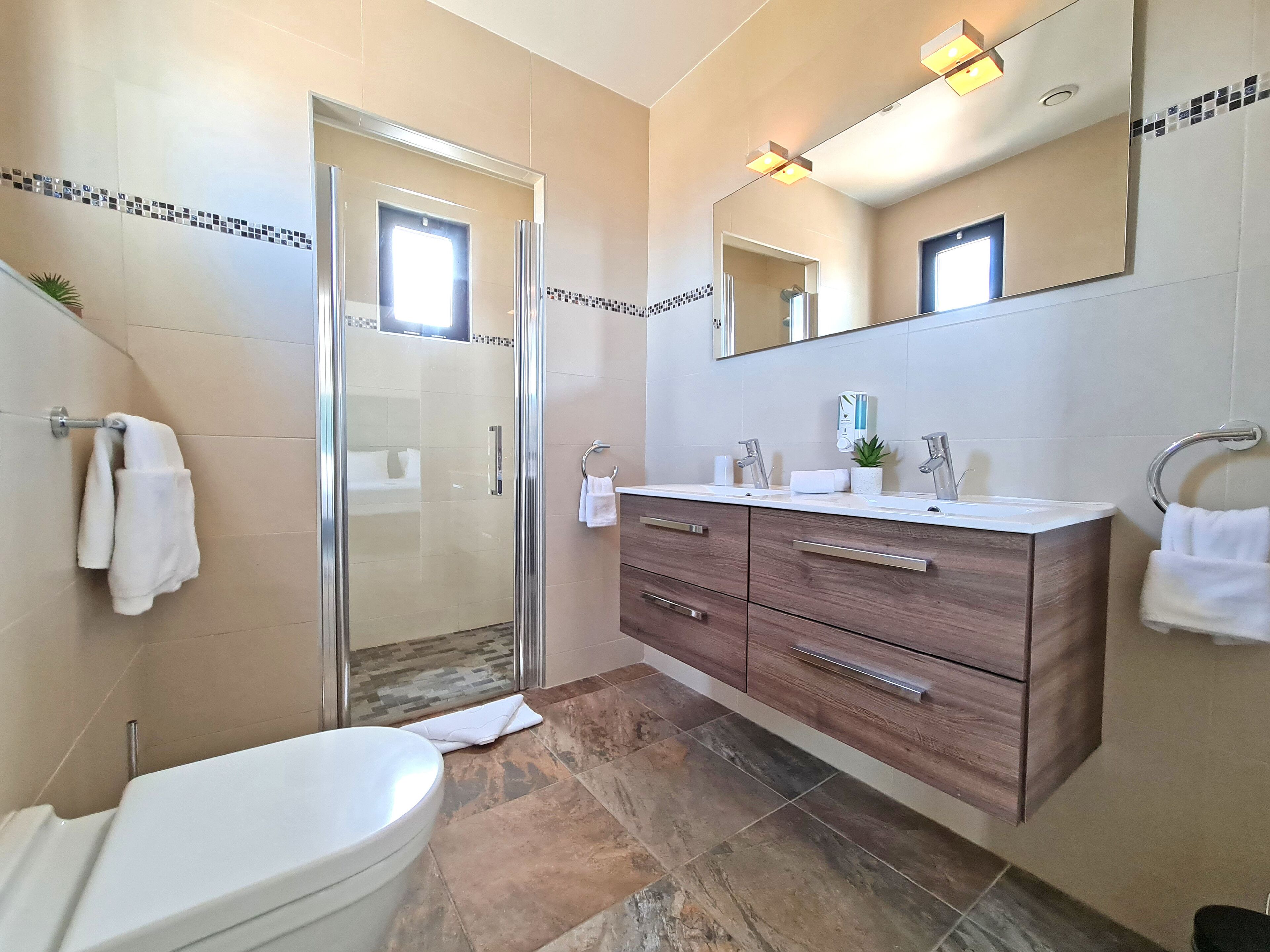 A modern bathroom with a glass shower enclosure, a white toilet, a wooden vanity with a mirror, and a sink with a towel rack.