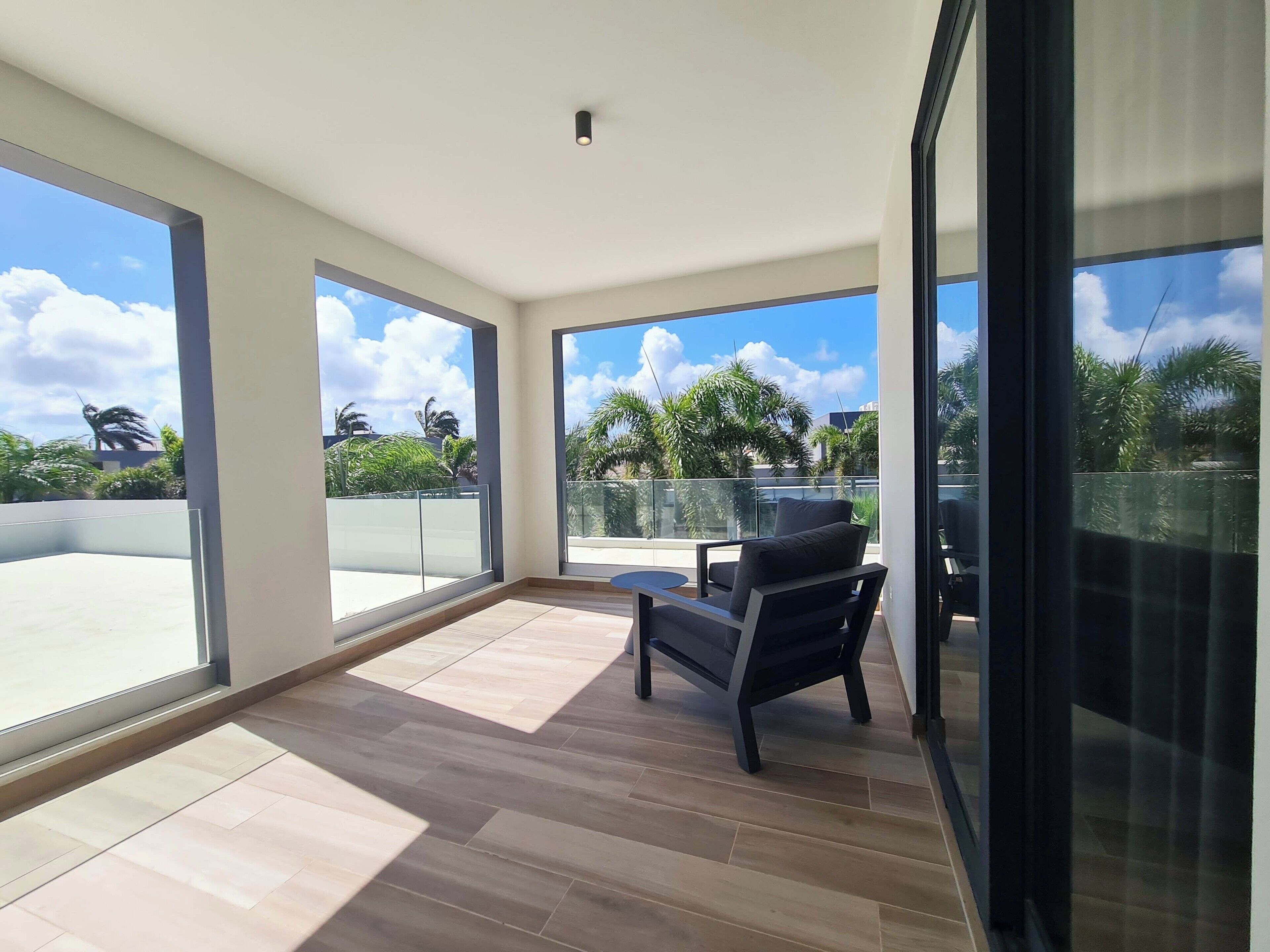 Un balcón amplio con ventanas grandes, una silla moderna y vistas a palmeras y un cielo despejado.