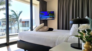 Habitación de hotel moderna con una cama grande, un televisor de pantalla plana en la pared, una mesita de noche con lámpara y vistas a palmeras a través de amplios ventanales.