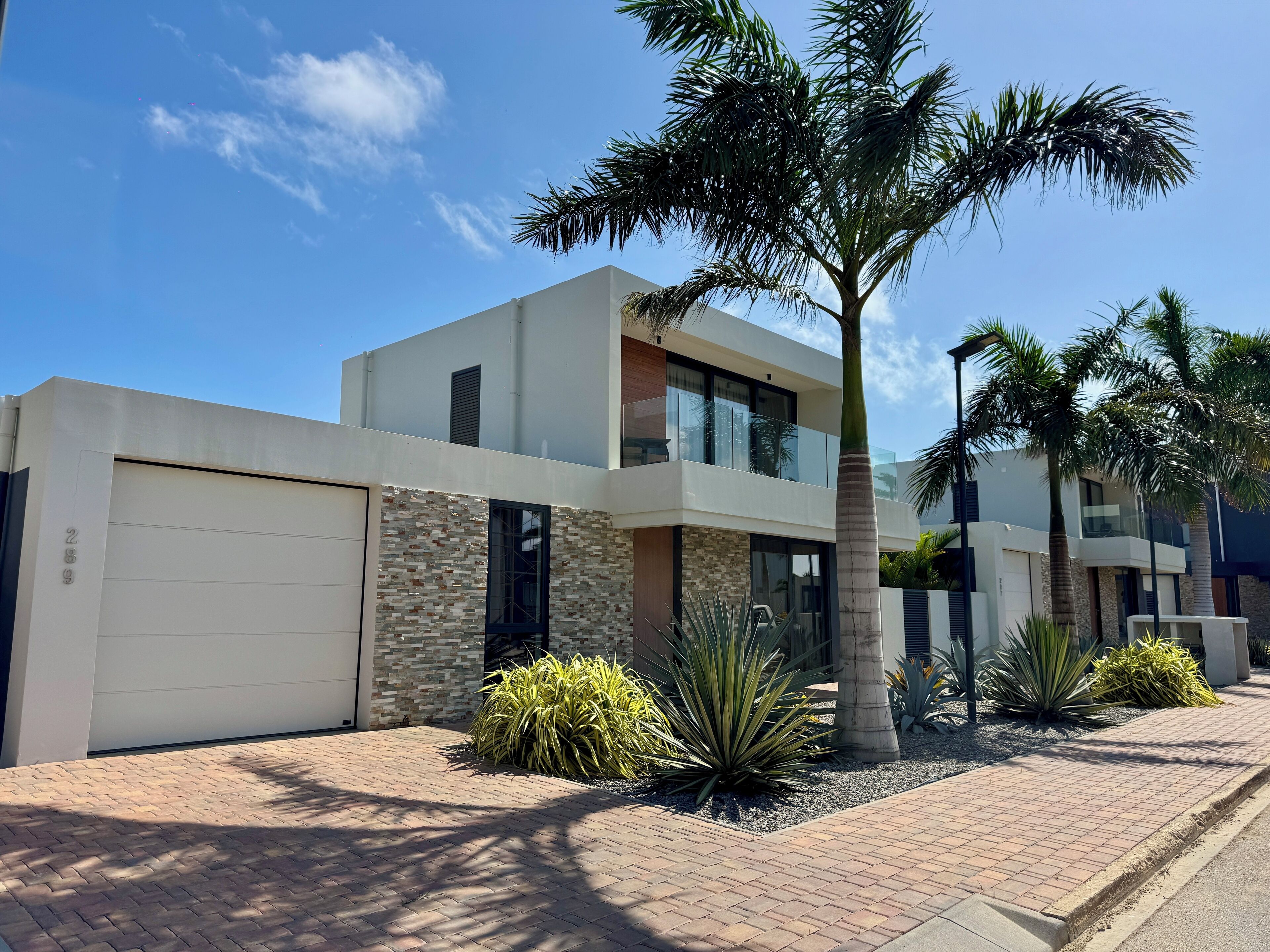 A modern two-story house with a garage, a balcony, and a well-maintained garden with palm trees and agave plants.