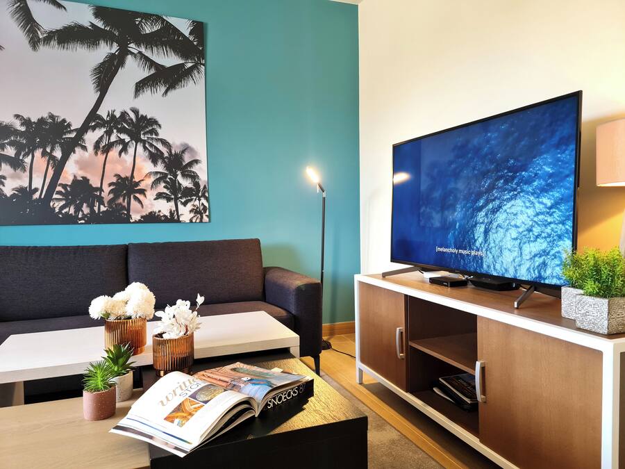 Una sala de estar con una pared color turquesa, un televisor de pantalla plana grande, un sofá, una mesa de centro con una revista y plantas, y una estantería.
