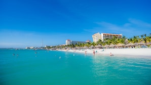 Una playa con un hotel al fondo, agua azul cristalina y palmeras.