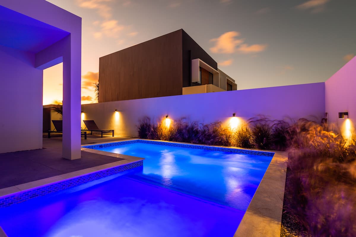 Un área de piscina exterior moderna con una piscina rectangular, rodeada por un muro bajo e iluminada con luces ambientales.