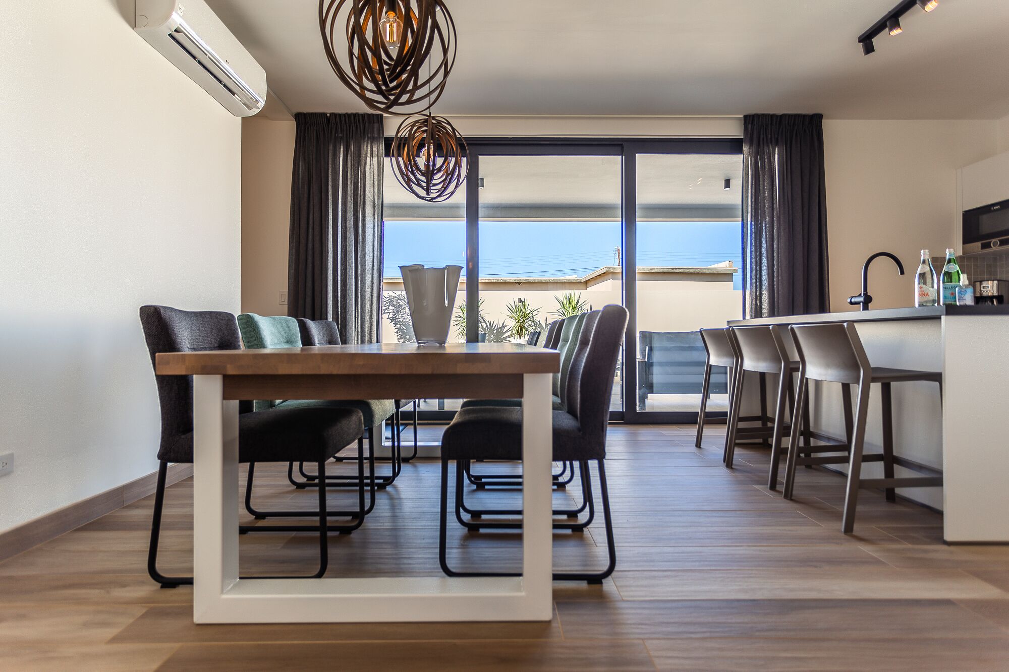 A modern dining area with a wooden table and chairs, a kitchen with a bar counter, and a large window with curtains.