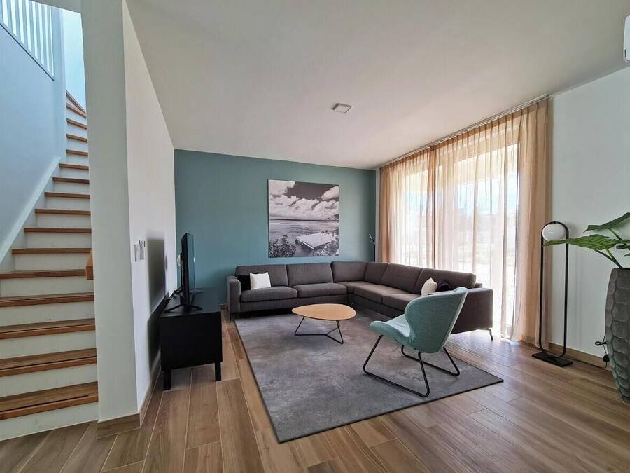 Una sala de estar moderna con un sofá modular, una mesa de centro y un televisor sobre un mueble bajo. Se observa una escalera a la izquierda y un ventanal con cortinas a la derecha.