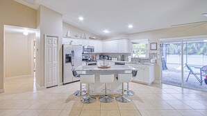 A modern kitchen with white cabinets, a central island with bar stools, and a view of a backyard through glass doors. - AMAZING HOUSE NEAR THE BEACHES OF SOUTH FLORIDA (Cape Coral)