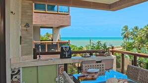 A balcony with a view of the ocean, a dining area with wicker furniture, and a kitchen area with a coffee maker.