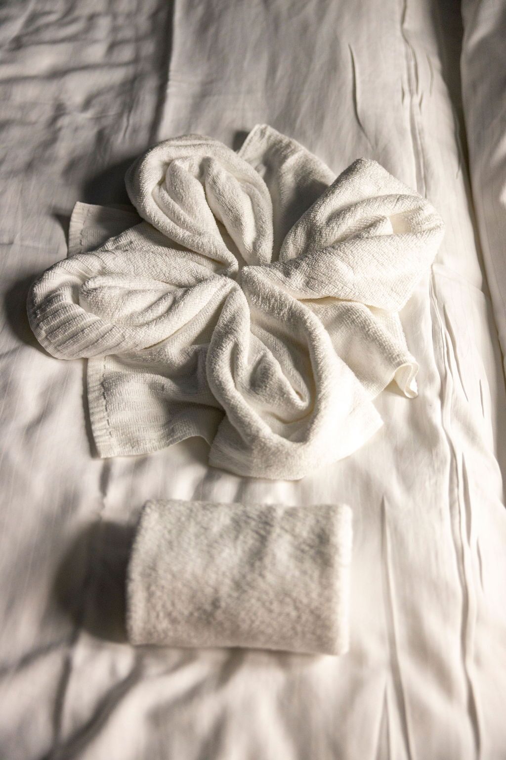 Una toalla blanca doblada con pulcritud y otra más pequeña sobre una cama con sábanas blancas texturizadas.
