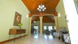 Un intérieur spacieux avec de hauts plafonds, des poutres en bois et un lustre. On y trouve un coin repas avec des chaises et une table, ainsi qu’un coin salon avec des poufs à motifs.