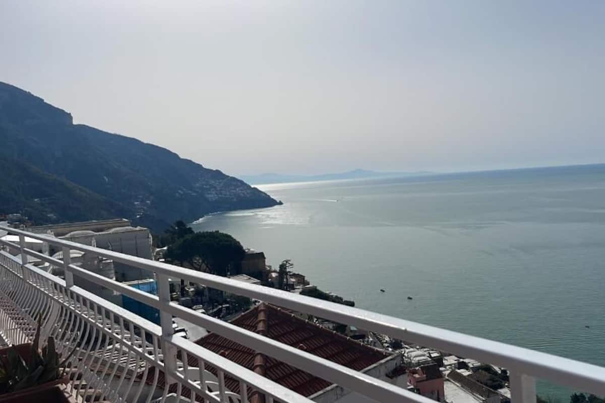 A coastal view from a balcony with a railing, overlooking a calm sea and distant mountains.