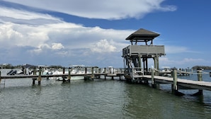 Un muelle con una torre de observación de madera, barcos amarrados y una masa de agua tranquila bajo un cielo parcialmente nublado.