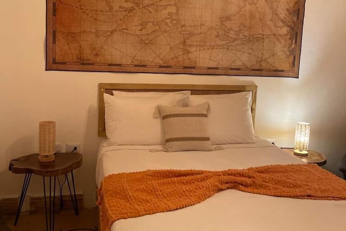 A hotel room with a large map artwork on the wall, a bed with white linens, an orange throw blanket, a small side table with a lamp, and a patterned rug.
