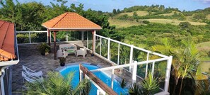 A rooftop pool area with a covered seating area, surrounded by lush greenery and hills in the background.